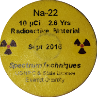  A disc source (also known as a button source) containing a sample of radioactive Sodium 22. It consists of an inch-wide plastic disc that is approximately an eighth of an inch thick. In the middle is a small sample of radioactive material, sealed in with epoxy.  The front face of the disc contains a sticker with information about the isotope, the date it was created, and the manufacturer. 